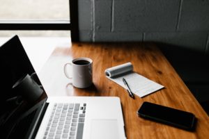 A desk with a laptop, notepad, coffee cup, and glasses case