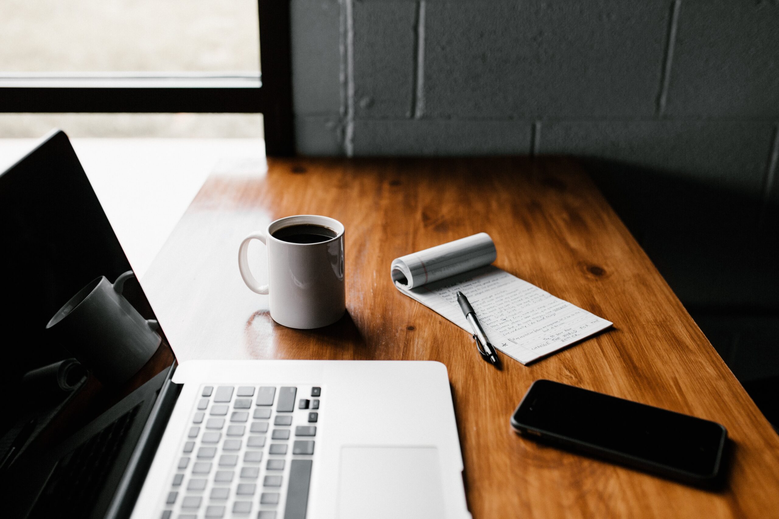 A desk with a laptop, notepad, coffee cup, and glasses case
