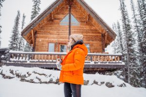 Woman in an orange winter jacket standing in snow outside a rustic cabin in the woods